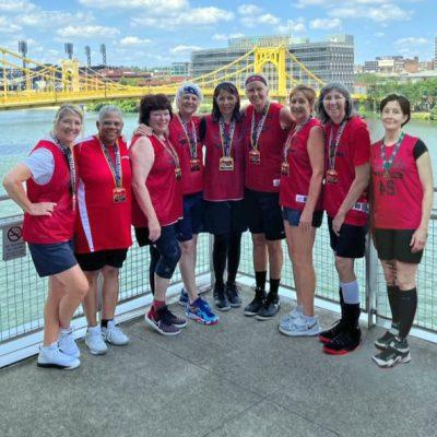 Nine older women in red and black basketball uniforms stand together with gold medals around their necks.